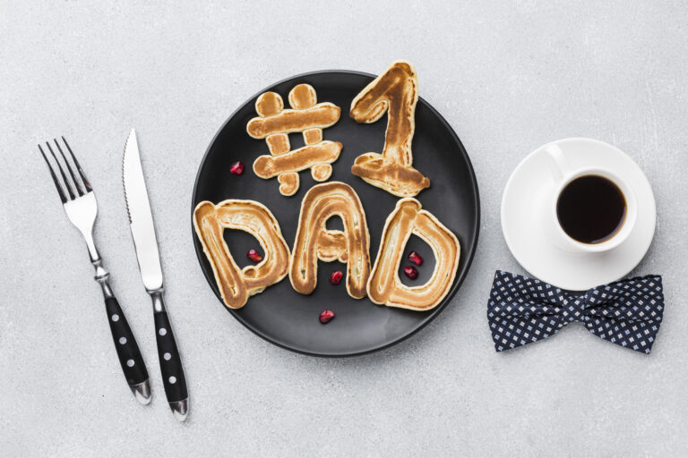 Image of a plate holding pancakes shaped to say "#1 dad", with cutlery to the left and a coffee cup to the right above a bow tie